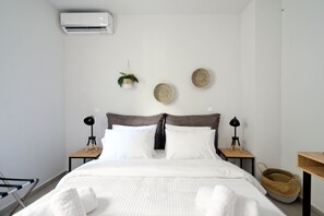 A neatly made bed with white linens, two bedside tables with lamps, a wall-mounted air conditioner, and decorative woven baskets.
