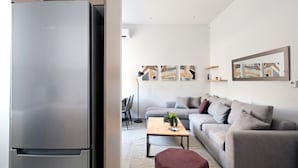 A modern living room with a grey sofa, a wooden coffee table, and a red ottoman. There is a large flat-screen TV mounted on the wall, and a refrigerator is visible in the foreground.