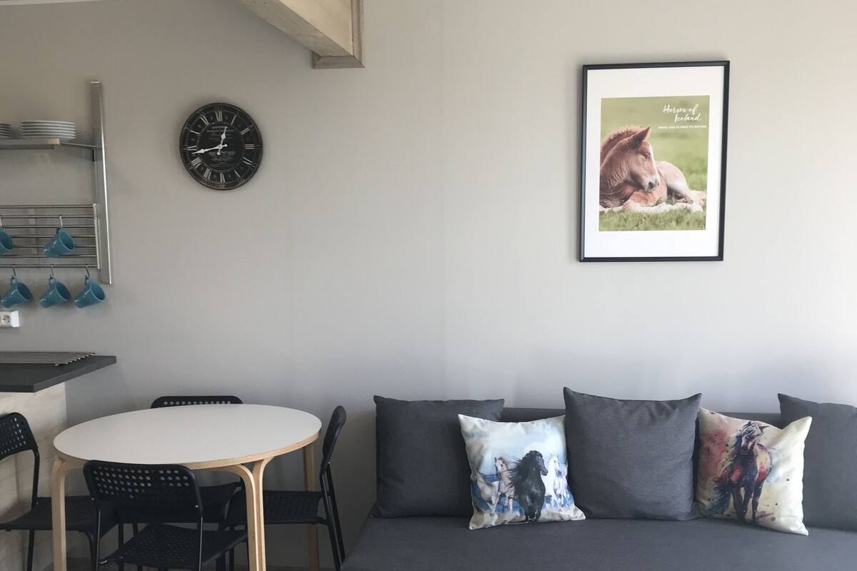 Un salon moderne avec un canapé gris, une table ronde et des chaises noires. Une horloge et un tableau représentant un cheval sont accrochés au mur.