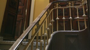A wooden staircase with a handrail, a door with a sign, and a wall-mounted light fixture. - Primavera (Amadora)