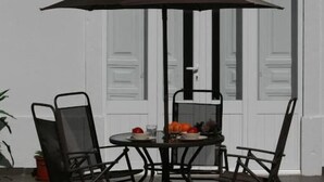 A round metal table with a dark umbrella, surrounded by four metal chairs, in front of a white door with glass panels. - Primavera (Amadora)