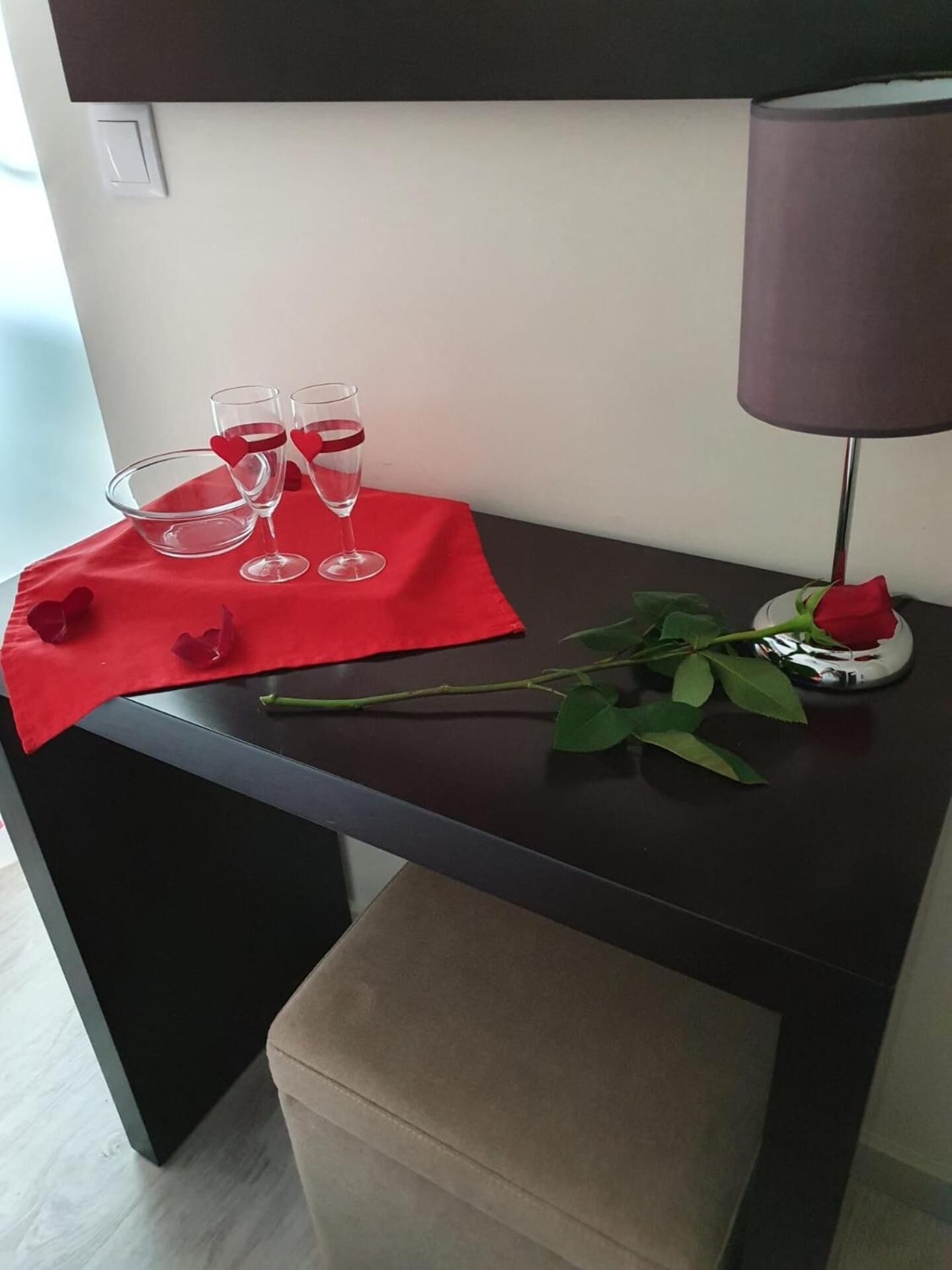 Een kleine tafel met een rode tafelkleed, twee wijnglazen, een roos en een lamp.