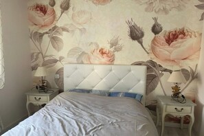 Ein Schlafzimmer mit einer gesteppten Kopfteil, einem Bett mit weißer und blauer Bettdecke, zwei Nachttischen mit Lampen sowie floralem Wandpapier.