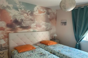 A bedroom with a tufted headboard, two beds with floral bedding, an orange pillow, a nightstand with a lamp, and a wall mural of clouds. - VILLA Biscarosse Ville - 6 pers. - 3 rooms (Biscarrosse)