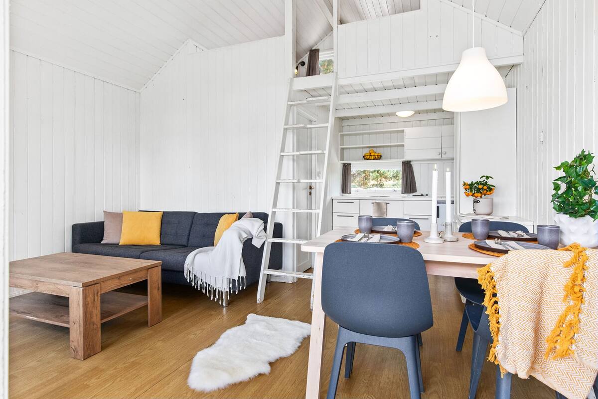 Een moderne, goed verlichte kamer met een houten salontafel, een bank met kussens, een eettafel met stoelen, en een witte trap die naar een hoger niveau leidt.