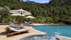 Zona de piscina con sillas de descanso y sombrillas, rodeada por una ladera boscosa.