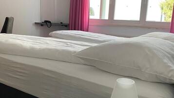 Una habitación de hotel con cama, una mesita de noche con lámpara, un televisor montado en la pared y una ventana con cortinas.