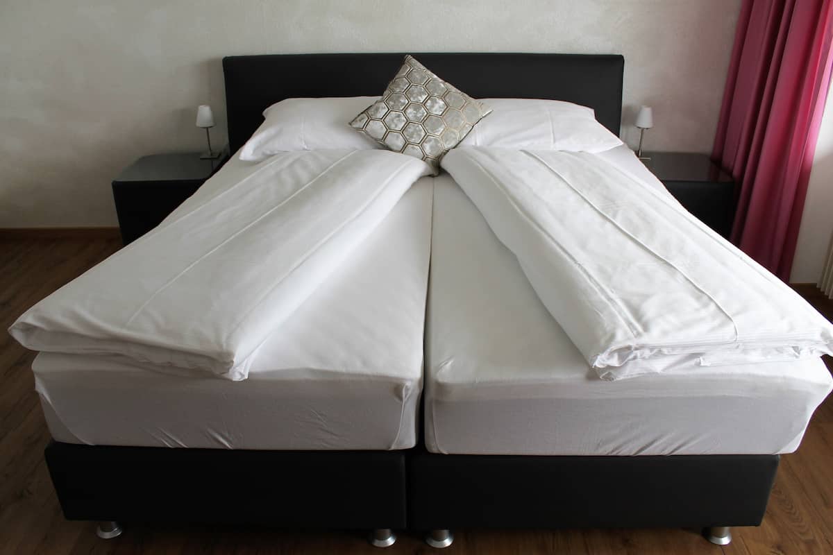 Un lit double avec du linge de lit blanc et un coussin décoratif, flanqué de deux tables de chevet avec des lampes, dans une pièce au plancher en bois et dotée d’une tête de lit sombre.