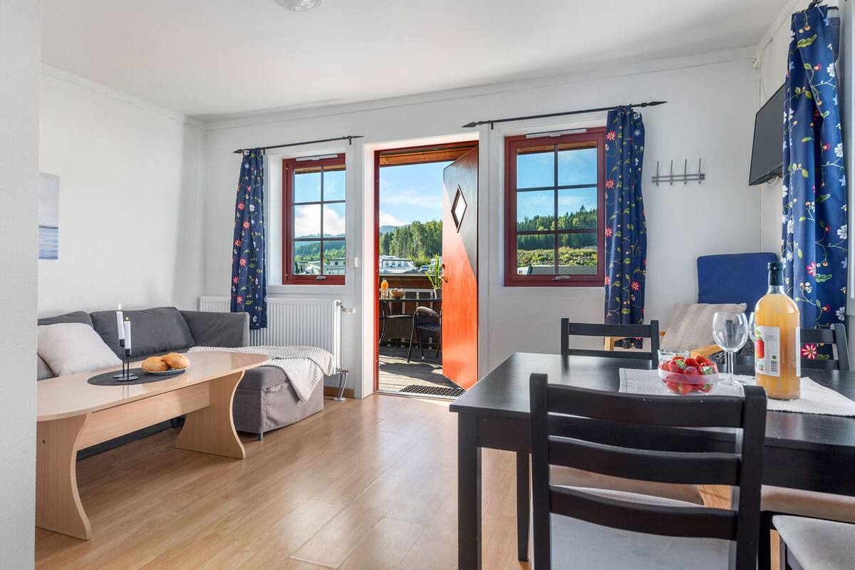 A modern living room with a sofa, a dining table, and a view of a garden through the open door.