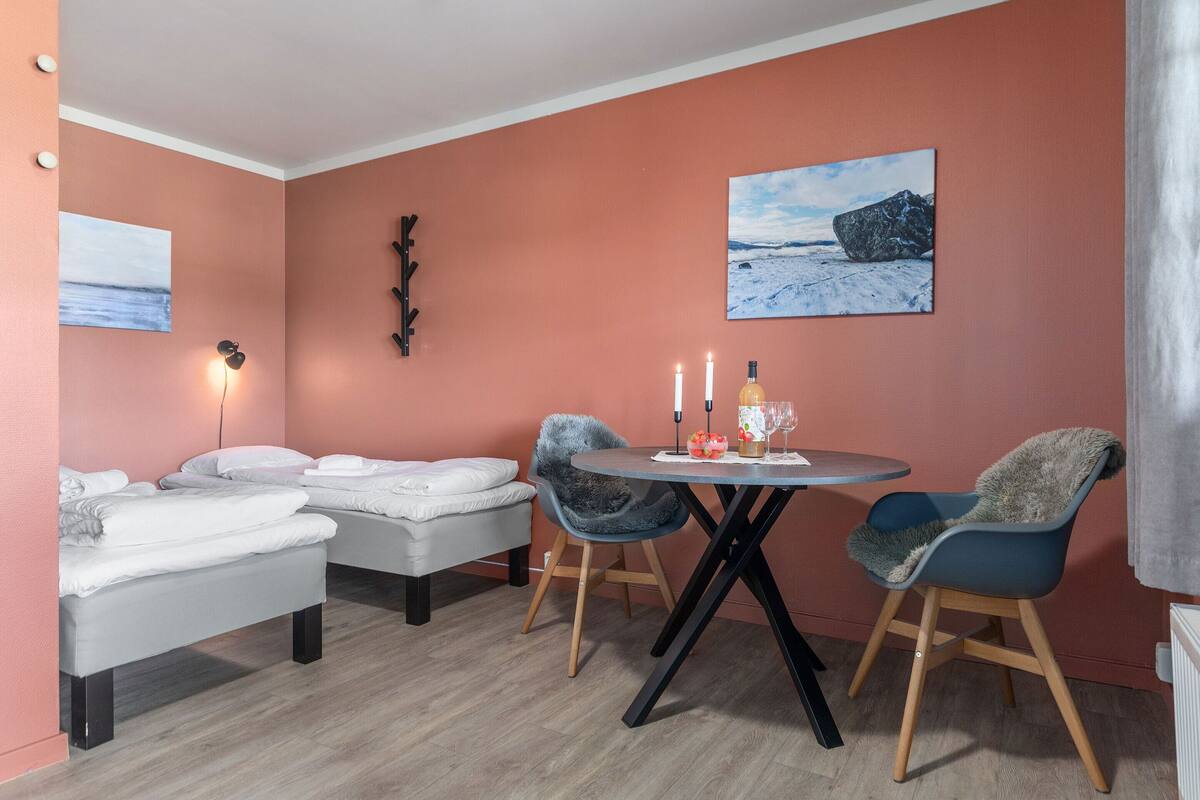 A room with two beds, a round table with chairs, a wall-mounted light, and two paintings.