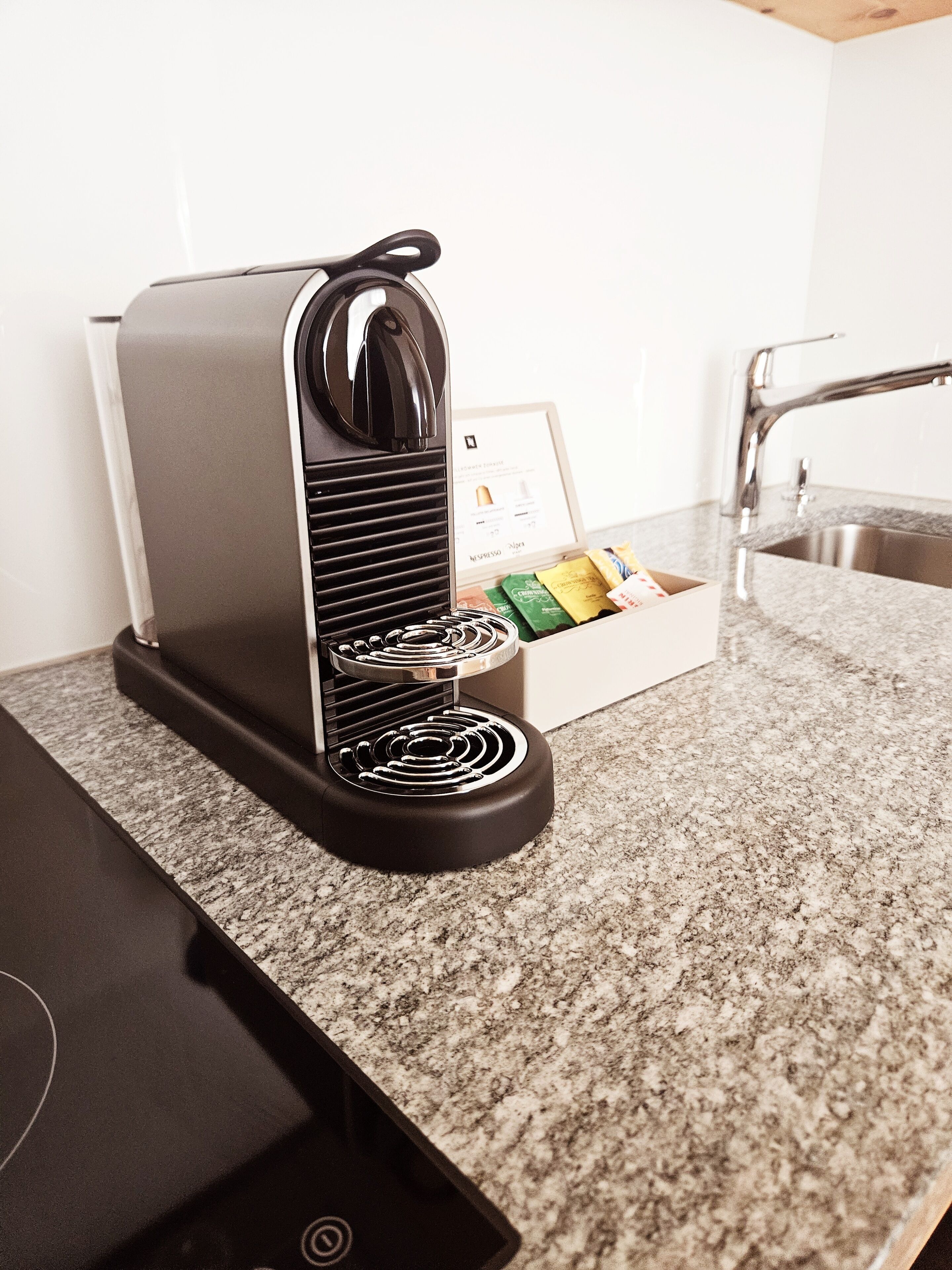 A kitchen with a coffee machine, a box of coffee pods, and a stainless steel sink.