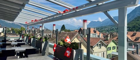 Una terraza con mobiliario exterior, banderas suizas y vistas a la montaña.