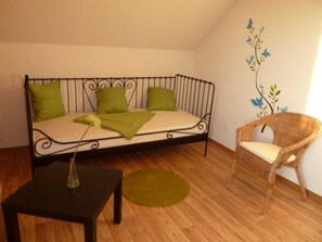 A small, cozy room with a metal-framed sofa, a wicker chair, a small table, and a green rug.