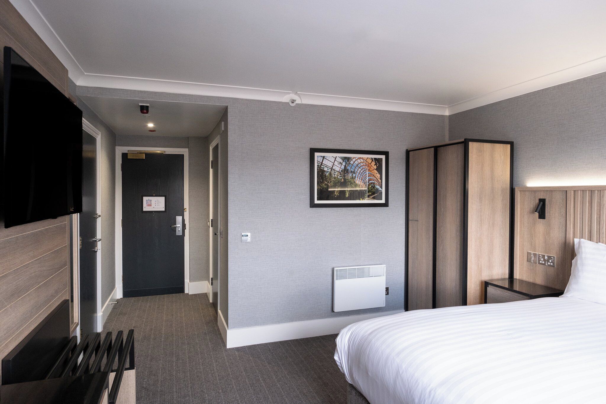 A modern hotel room with a bed, a flat-screen TV, a wardrobe, and a framed picture on the wall.