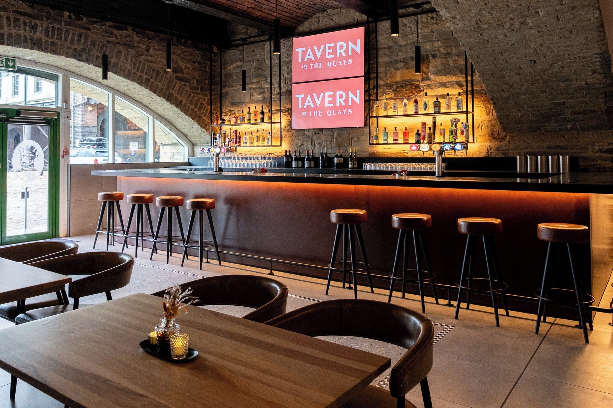 Una zona de bar con taburetes, una barra llena de botellas y un cartel que dice "TAVERN ON THE QUAYS".