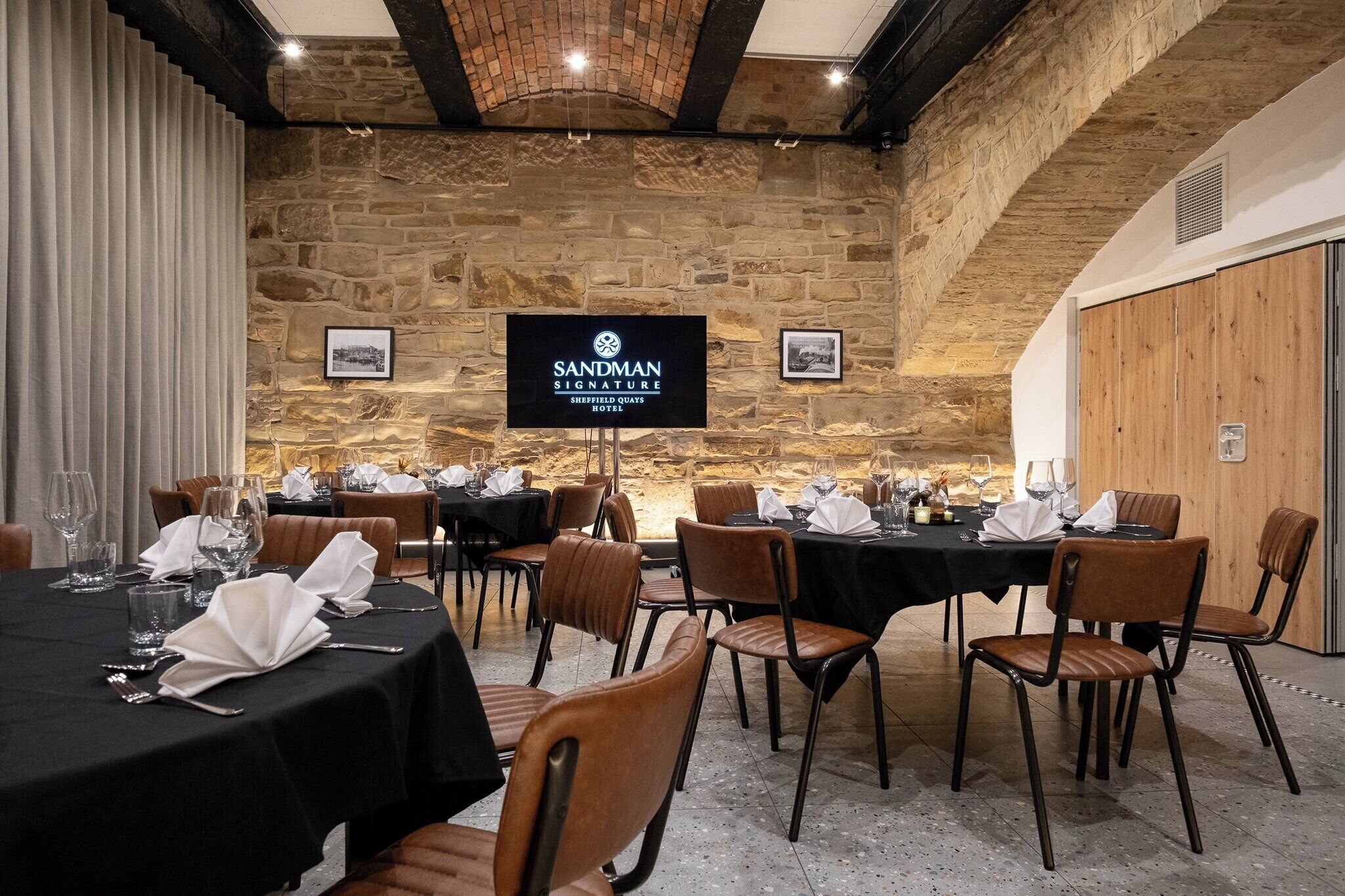 A conference room with multiple tables set for dining, stone walls, and a sign for Sandman Signature Sheffield Quays Hotel.