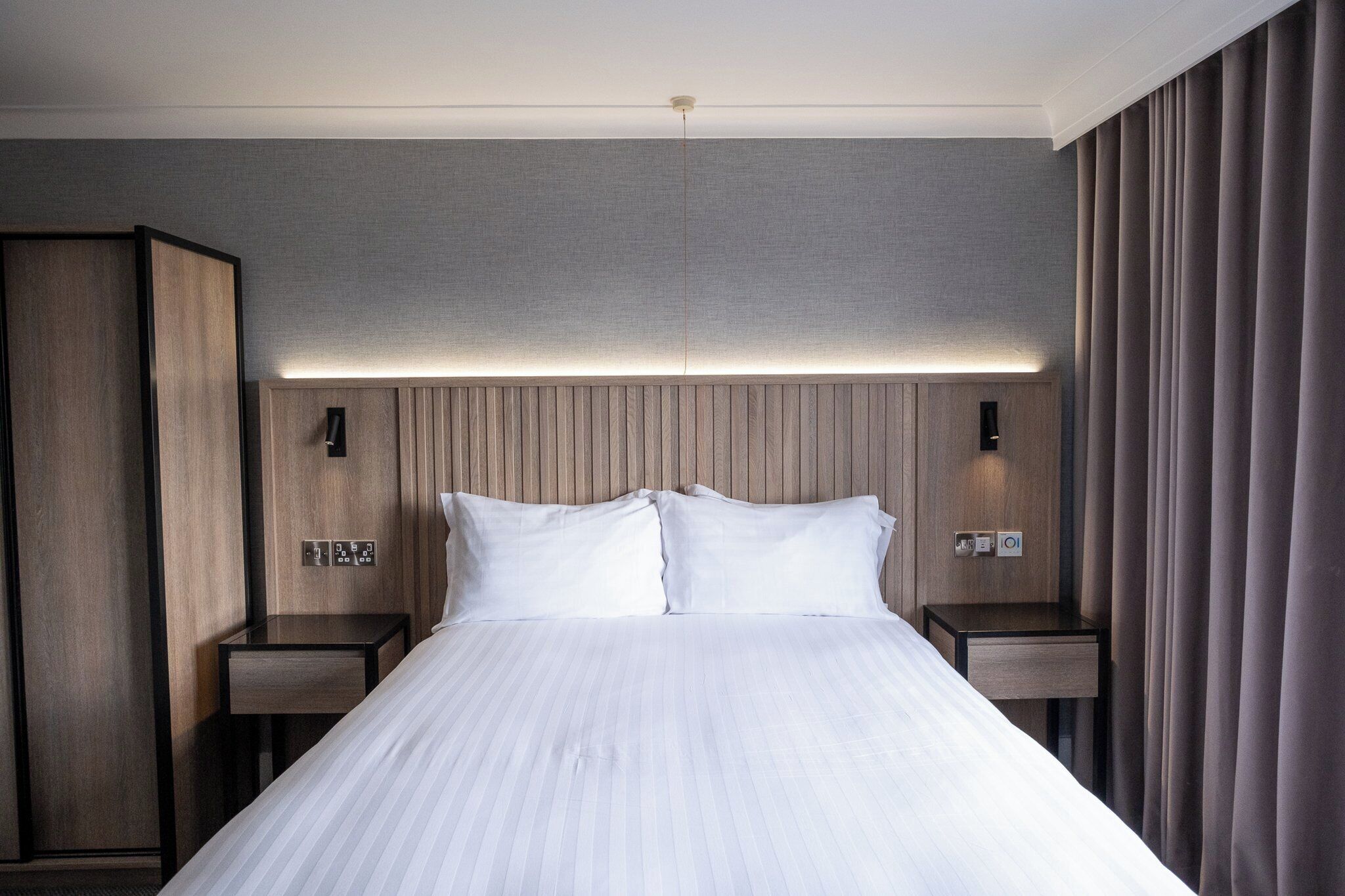 A hotel room with a large bed, two bedside tables, a wardrobe, and a headboard with integrated lighting.