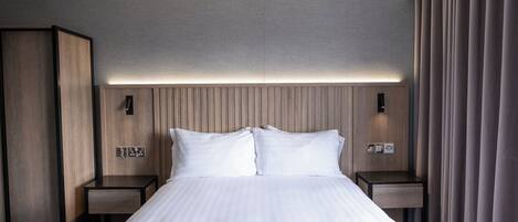 A hotel room with a large bed, two bedside tables, a wardrobe, and a headboard with integrated lighting.