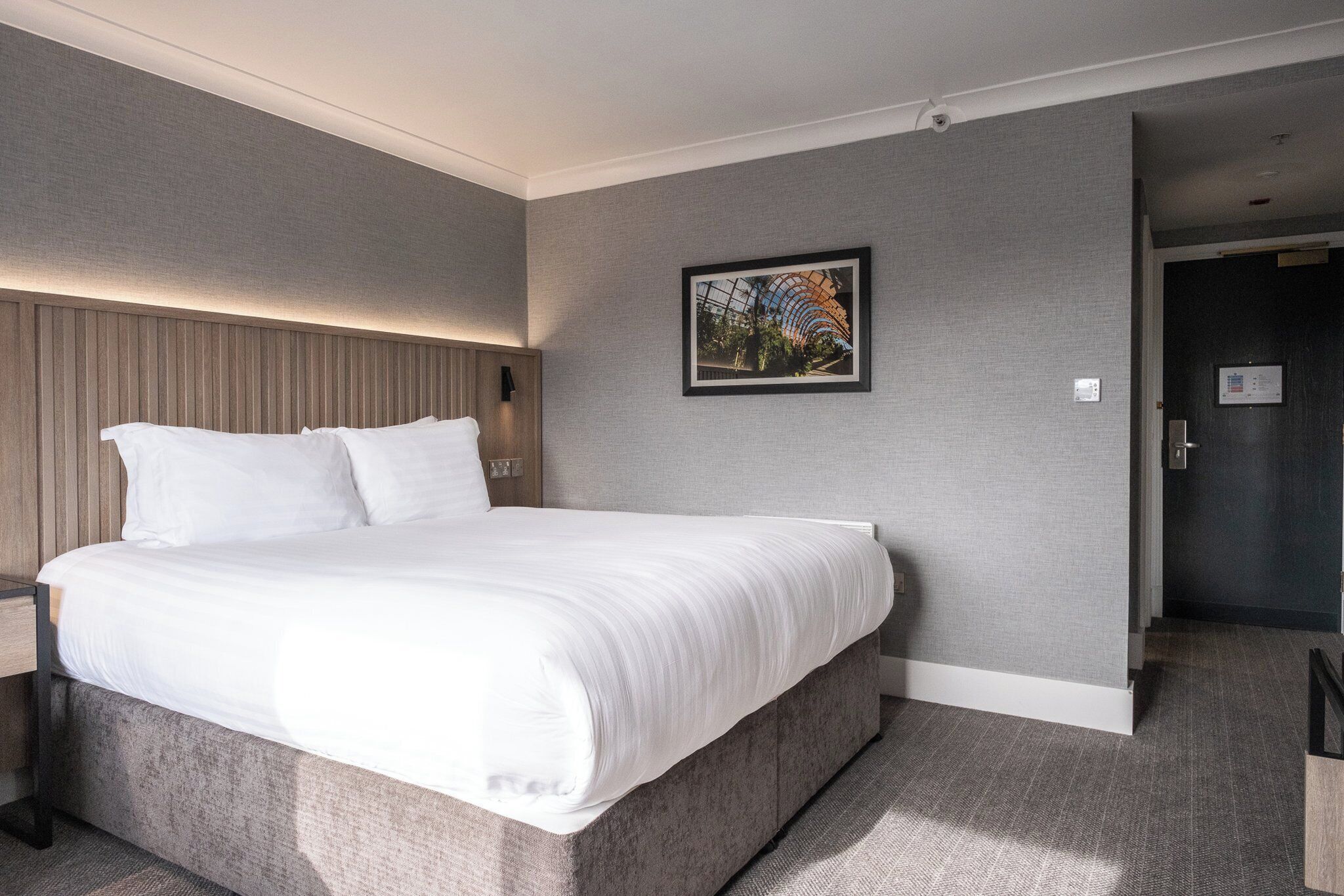 A modern hotel room with a large bed, a headboard with vertical lines, a framed picture on the wall, and a door leading to another room.