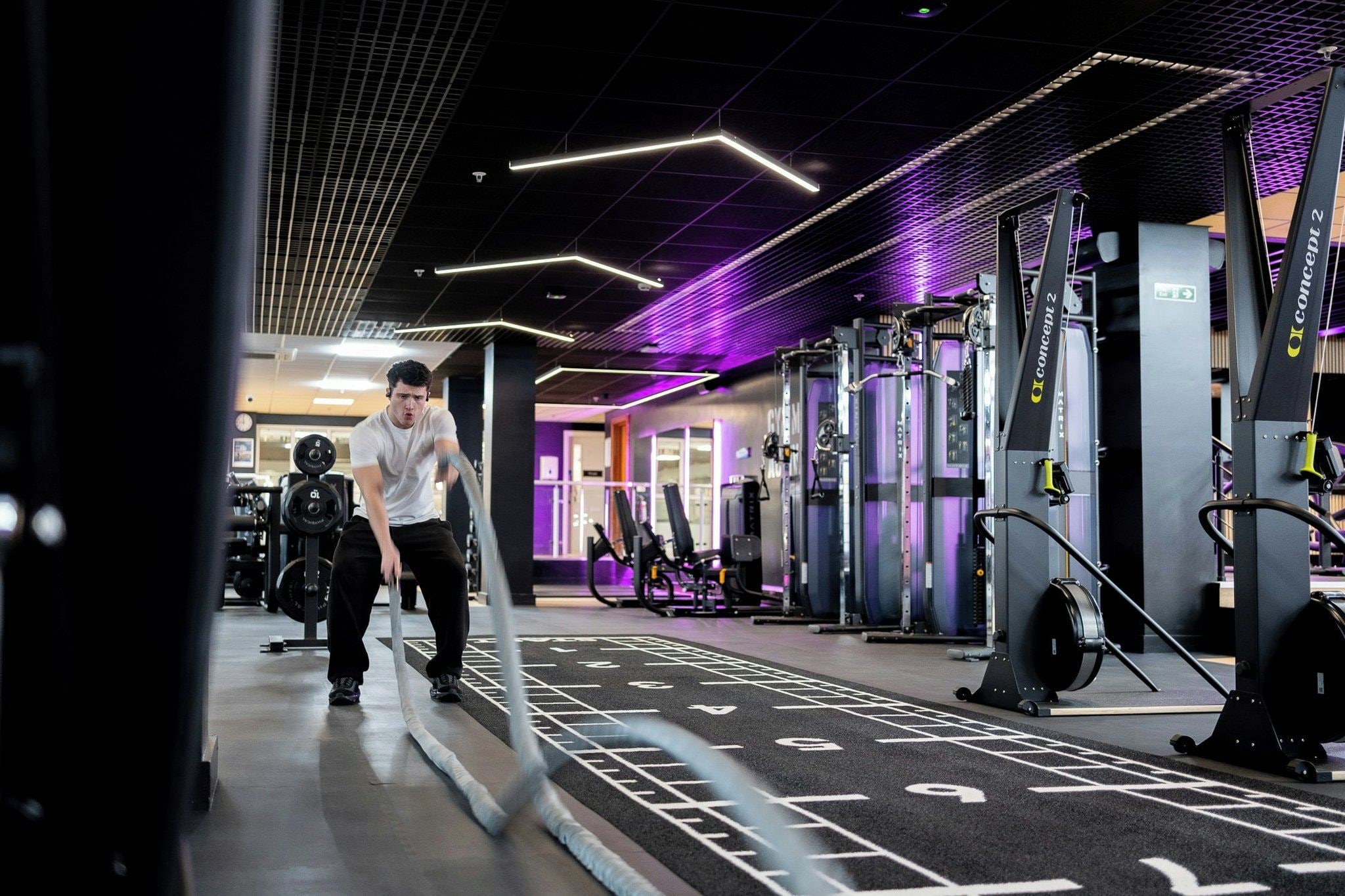 A gym with exercise equipment, a person running on a track, and a ceiling with geometric lighting.