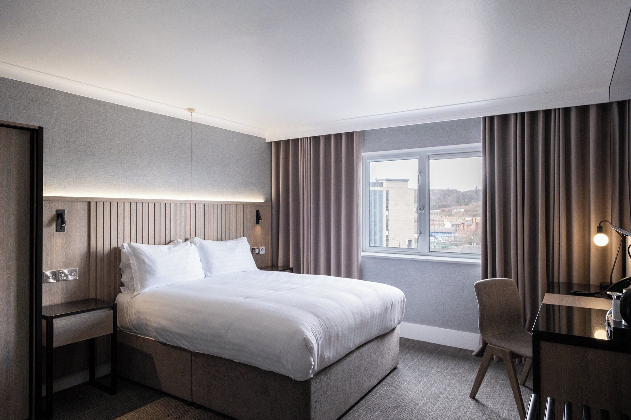 A modern hotel room with a large bed, a desk, and a view of the city through the window.