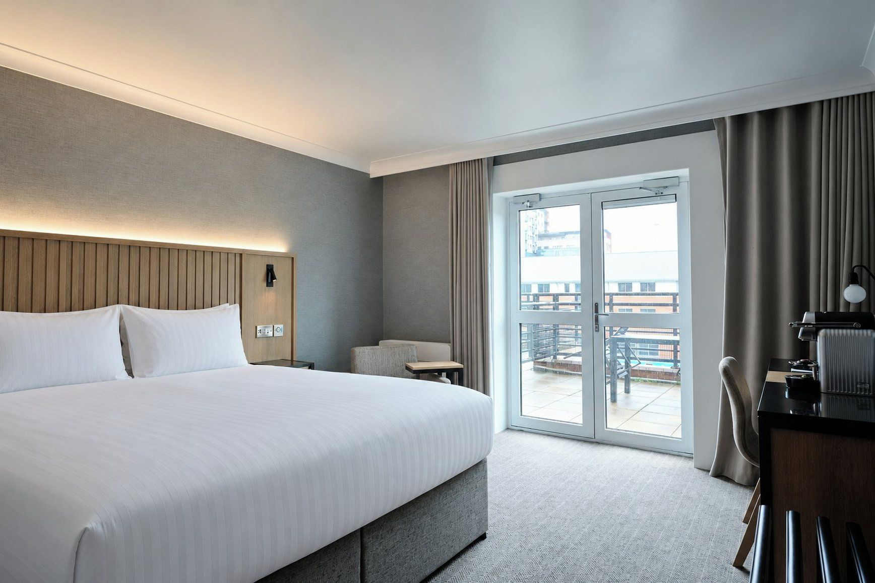 A hotel room with a large bed, a desk, a chair, and a view of the outdoors through a sliding door.