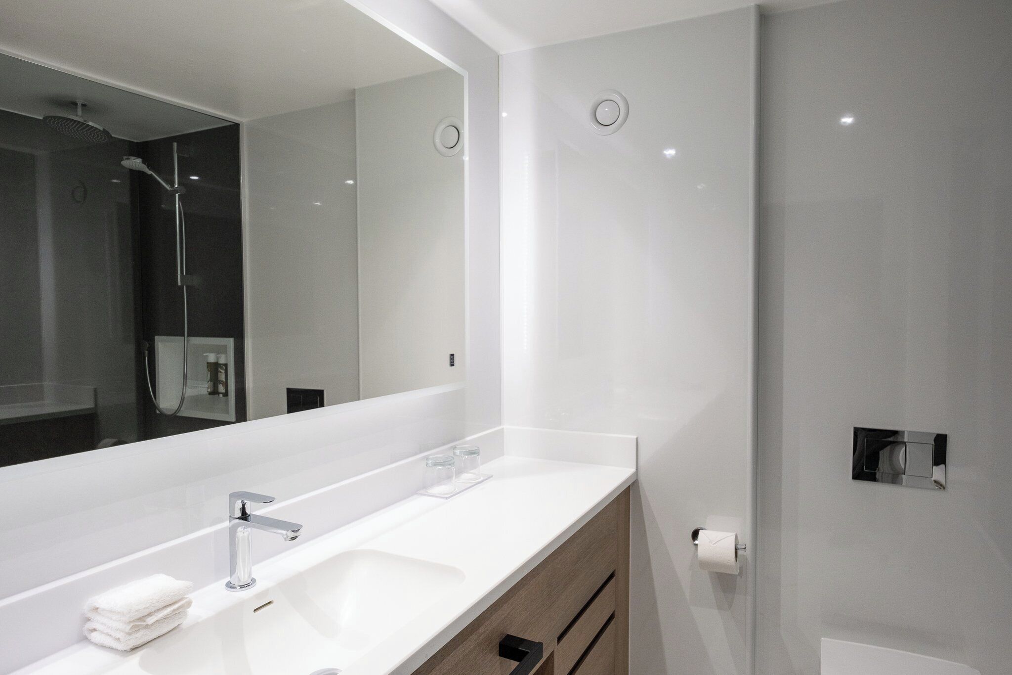 A modern bathroom with a large mirror, two sinks, a shower, and a toilet paper holder.