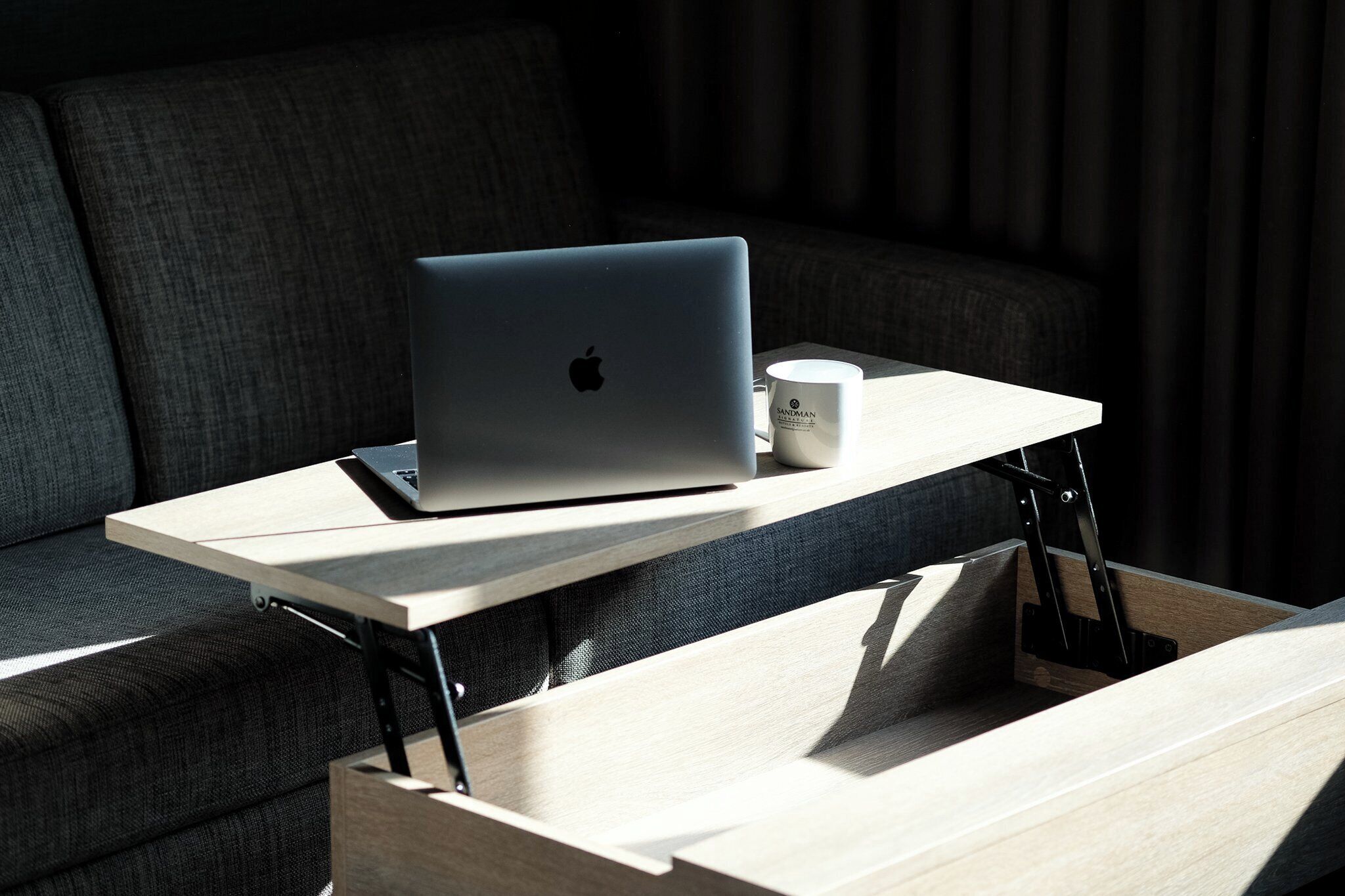 A laptop on a wooden desk with a white mug beside it, placed on a couch with a dark fabric.