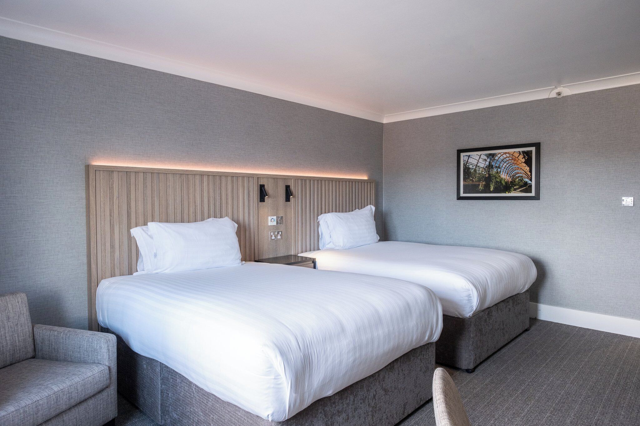 Une chambre d’hôtel avec deux lits, un canapé et une tête de lit dotée d’un éclairage intégré.