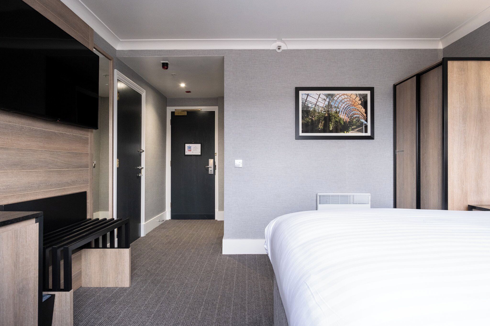 Una habitación de hotel con una cama, un televisor montado en la pared, una puerta que da a un pasillo y un cuadro enmarcado.
