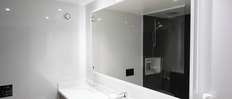 A modern bathroom with a large mirror, white vanity, and a shower visible in the reflection.