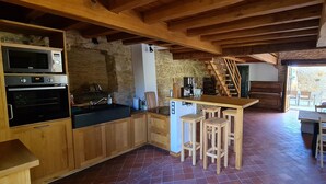 Eine rustikale Küche mit Holzsäulen, einem eingebauten Ofen, einer Mikrowelle und einer Kaffeemaschine. Es gibt eine Holzbar mit Hockern und eine Treppe, die zu einer oberen Ebene führt.