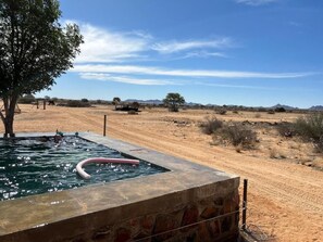 Una piscina con borde de piedra, rodeada por un paisaje seco y abierto con vegetación escasa y montañas al fondo.