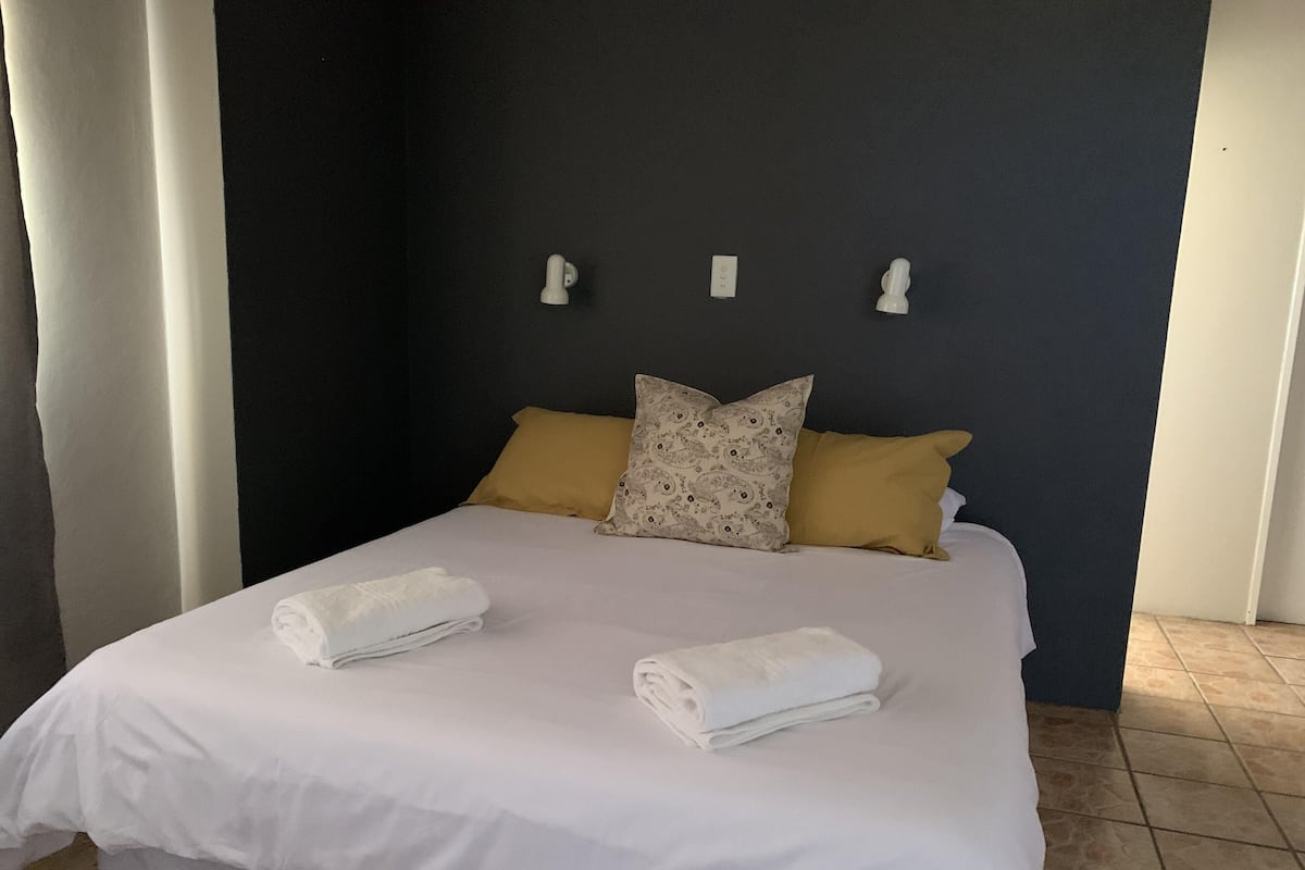 Cama bien hecha con sábanas blancas, dos toallas enrolladas, pared oscura y dos almohadas, una amarilla y otra con diseño estampado.