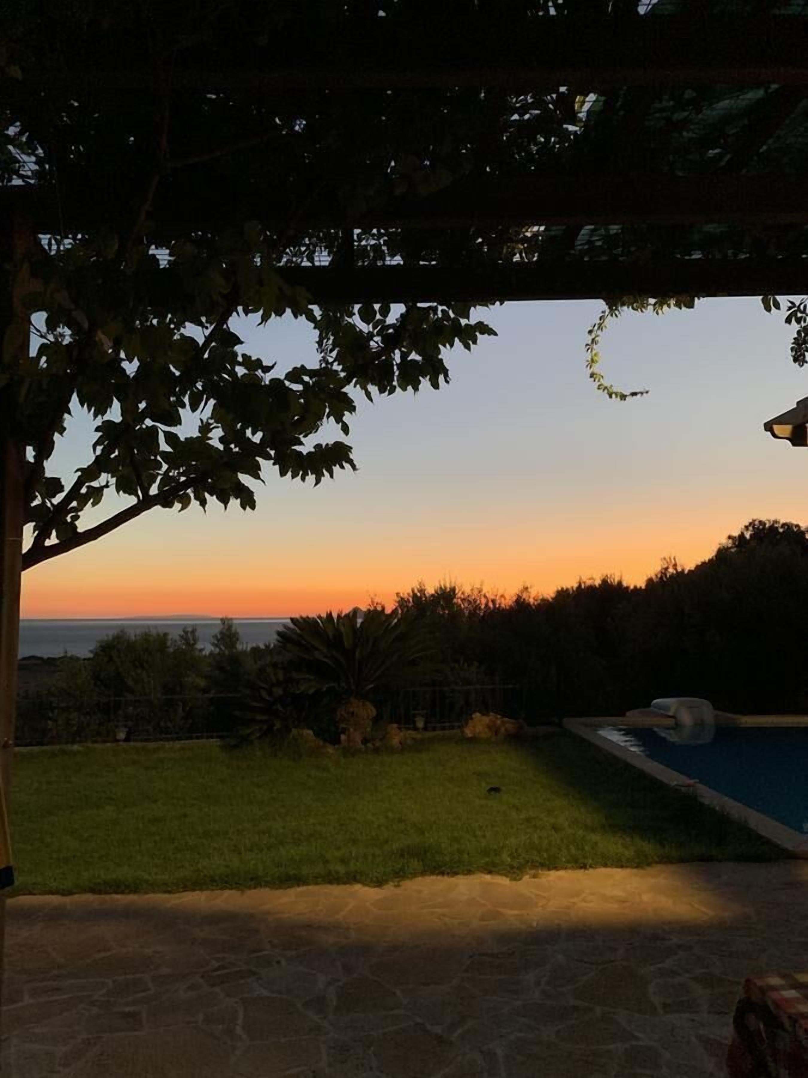 A patio area with a view of the ocean at sunset, a swimming pool, and a stone-tiled floor.