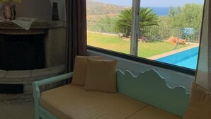 A room with a sofa facing a large window overlooking a pool and greenery. - Villa Georgia (Faistos)