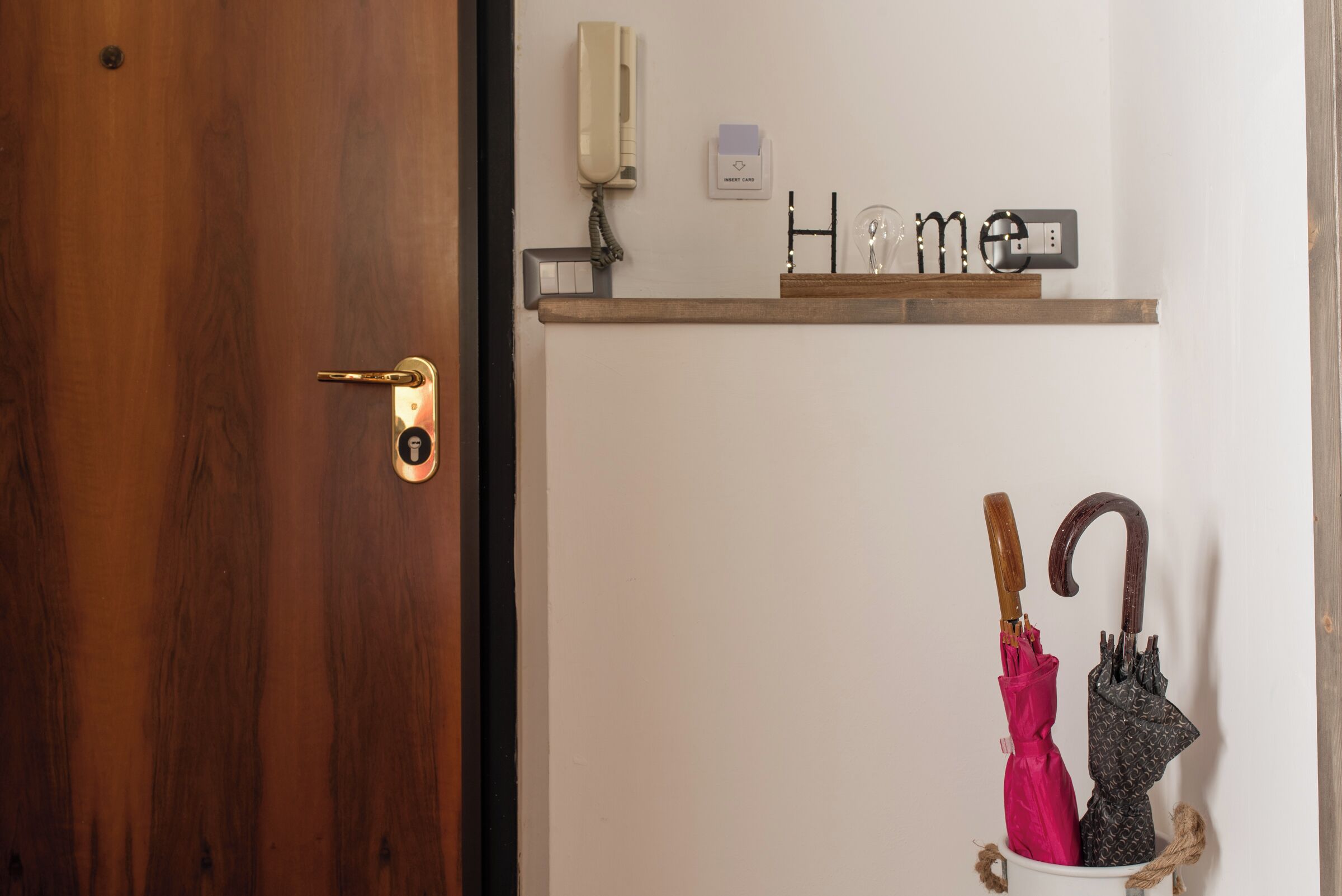 Une porte en bois avec une poignée en laiton, un interphone fixé au mur, une étagère sur laquelle est écrit le mot « Accueil » et un interrupteur, ainsi qu’un seau blanc contenant deux parapluies.