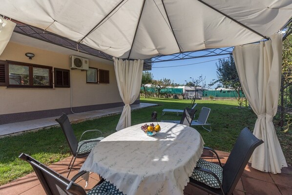 An outdoor patio area with a round table covered by a white tablecloth, surrounded by metal chairs with green cushions.