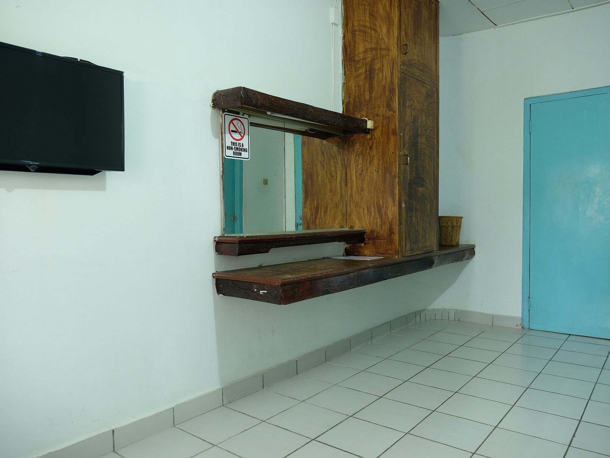 A room with a wooden door, a no-smoking sign, a TV mounted on the wall, and a tiled floor.
