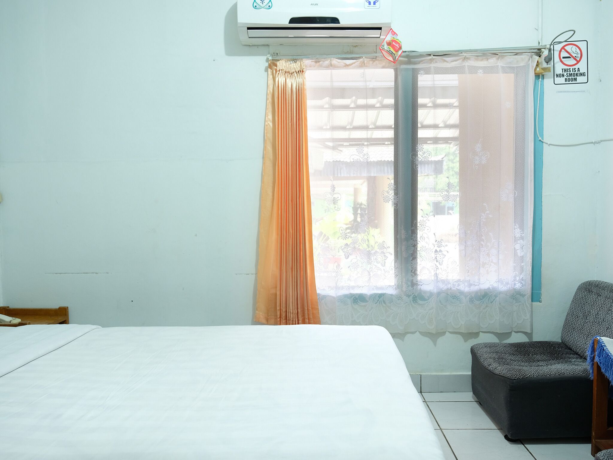 A hotel room with a bed, a chair, a window with curtains, an air conditioning unit, and a non-smoking sign.