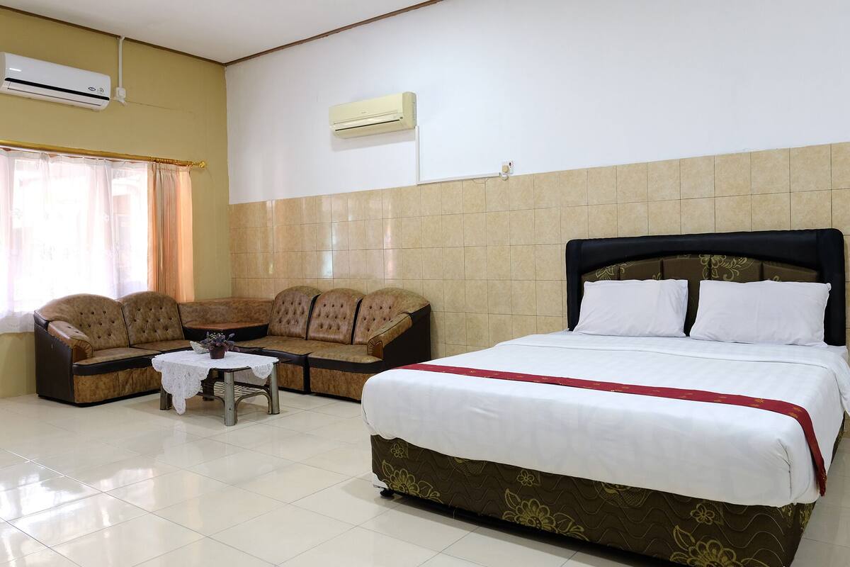 A hotel room with a bed, a sofa, a small table with a vase, and an air conditioning unit on the wall.