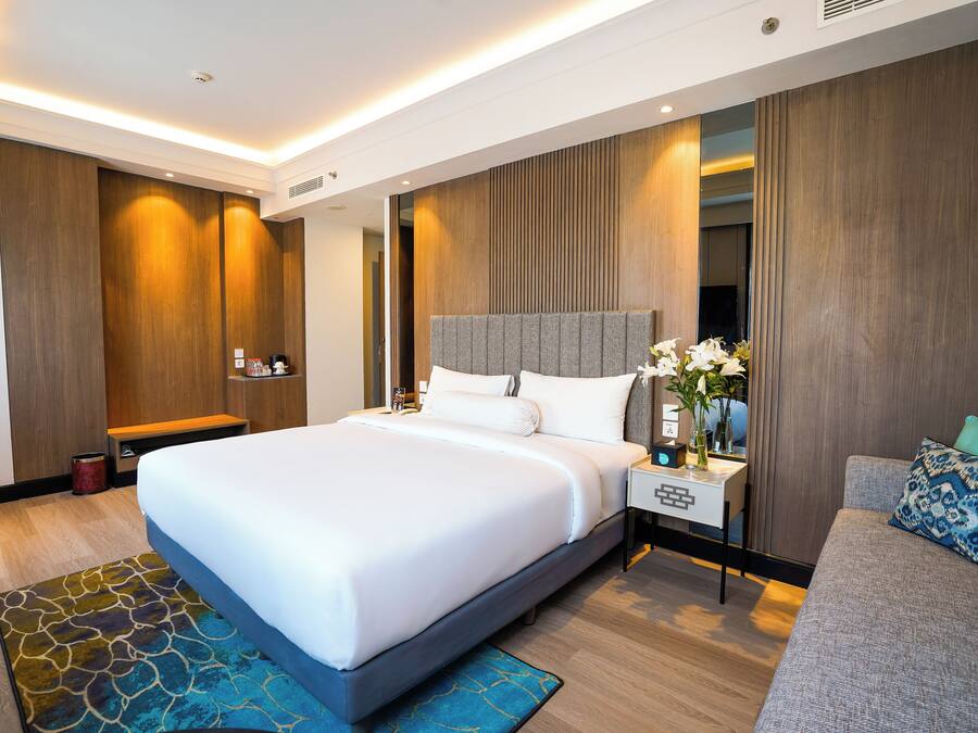 Una habitación de hotel moderna con una cama grande, un sofá gris, una mesita de noche con un florero y paneles de madera en las paredes.