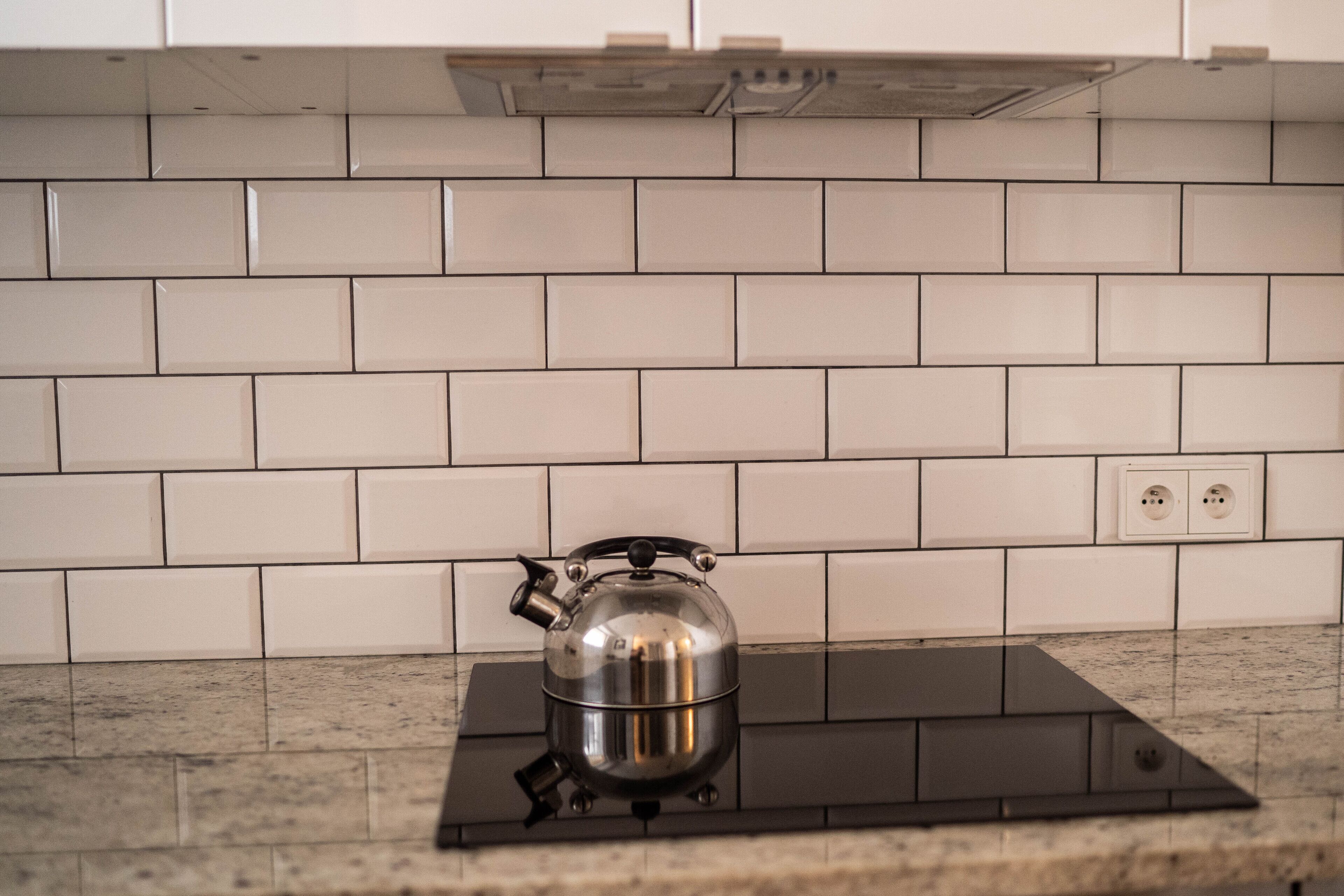 A kitchen with white subway tiles, a stainless steel kettle on an induction cooktop, and a double electrical outlet on the wall.