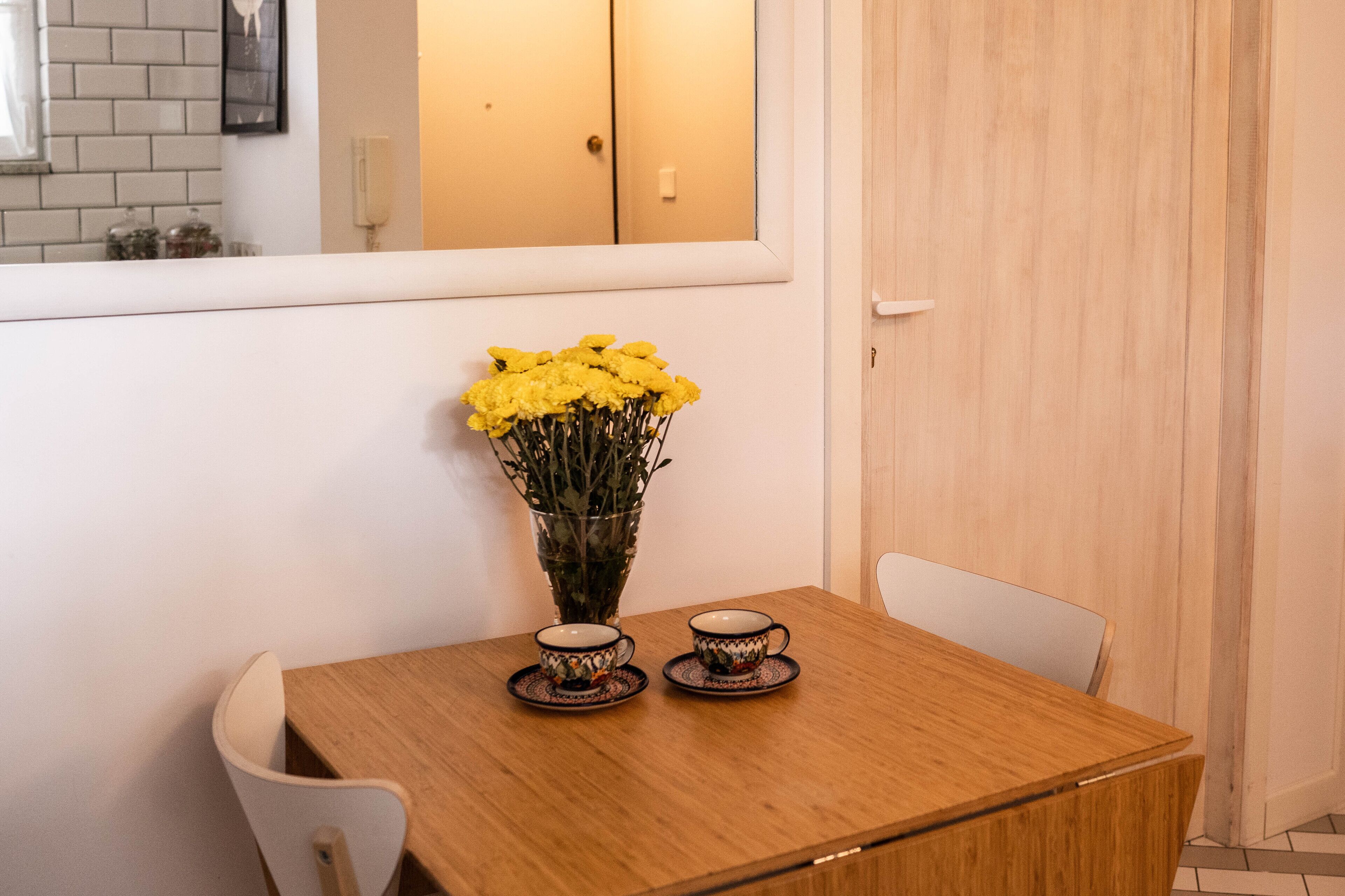 Una mesa de madera con dos tazas y platillos a juego, un florero con flores amarillas y una silla blanca.