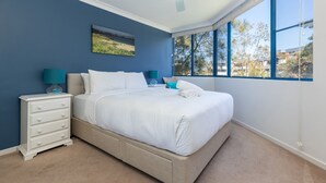 A bedroom with a bed, a nightstand, a lamp, and a window with a view of trees and buildings. - Enjoy wonderful water views from Heritage 303 (Tuncurry)