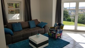 Ein Wohnzimmer mit einem braunen Sofa, einem farbenfrohen Sessel, einem weißen Couchtisch und Blick auf eine grüne Landschaft durch große Fenster.