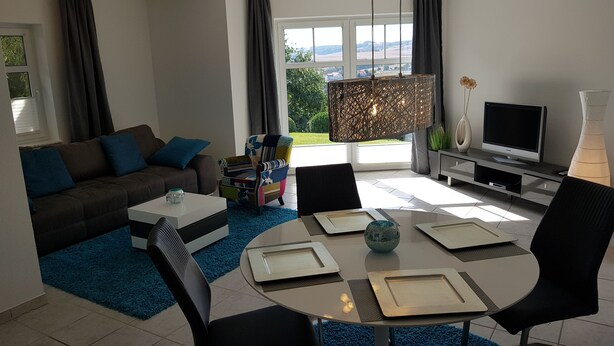 Ein modernes Wohnzimmer mit einer Couch, einem Couchtisch und Sesseln, einem Fernseher und einem großen Fenster mit Blick ins Freie.