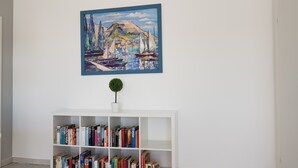 Ein Zimmer mit einem weißen Bücherregal, das mit Büchern gefüllt ist, einer kleinen Topfpflanze und einem farbenfrohen Gemälde einer Küstenlandschaft  über dem Bücherregal.
