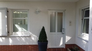 Ein Haus mit weißer Fassade, einem Backsteinfundament, einem Topf-Immergrün und einer Tür mit einem Milchglasfenster.