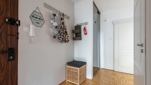 A hallway with a wooden door, a white wall with a hexagonal sign, a coat rack with a patterned bag, a small wooden storage unit, and a white door at the end. - P&O Apartments Okecie 5 (Warsaw)